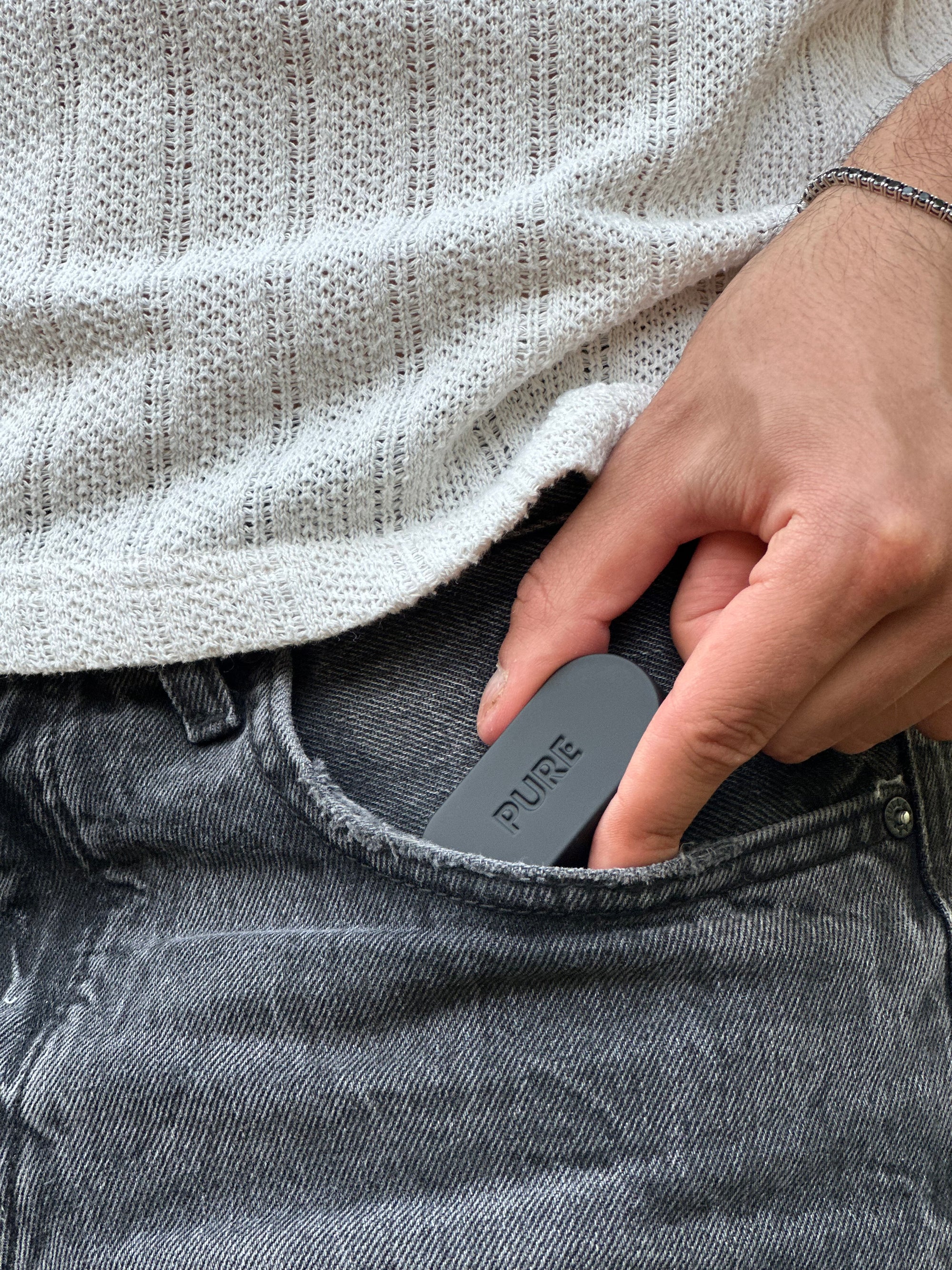 Close-up of a hand sliding the compact Pure solid perfume case into the front pocket of black denim jeans, highlighting its portable and pocket-sized design