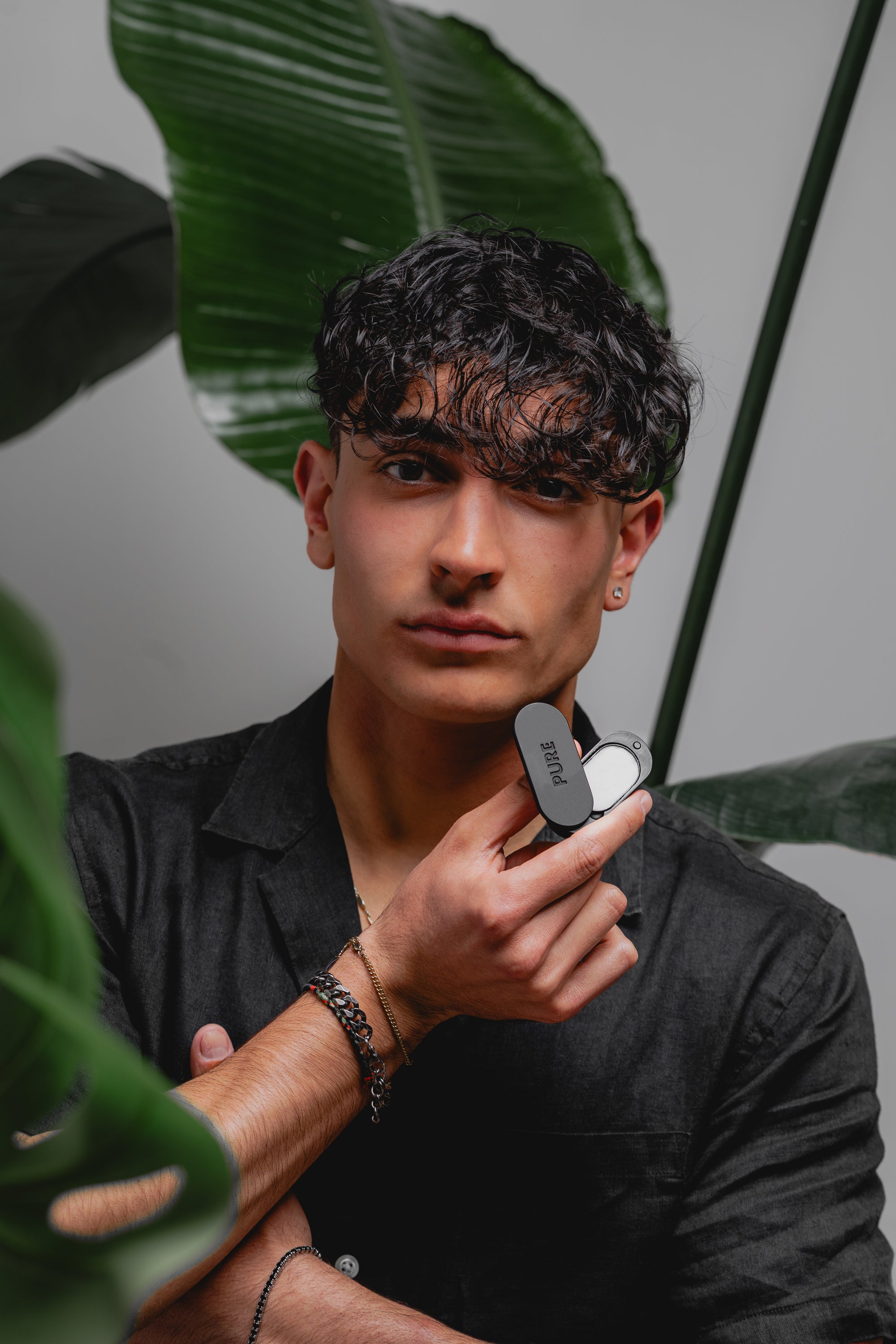 Man holding Pure solid perfume in a sleek black case, standing in front of tropical leaves, demonstrating the compact and stylish design of the fragrance product.